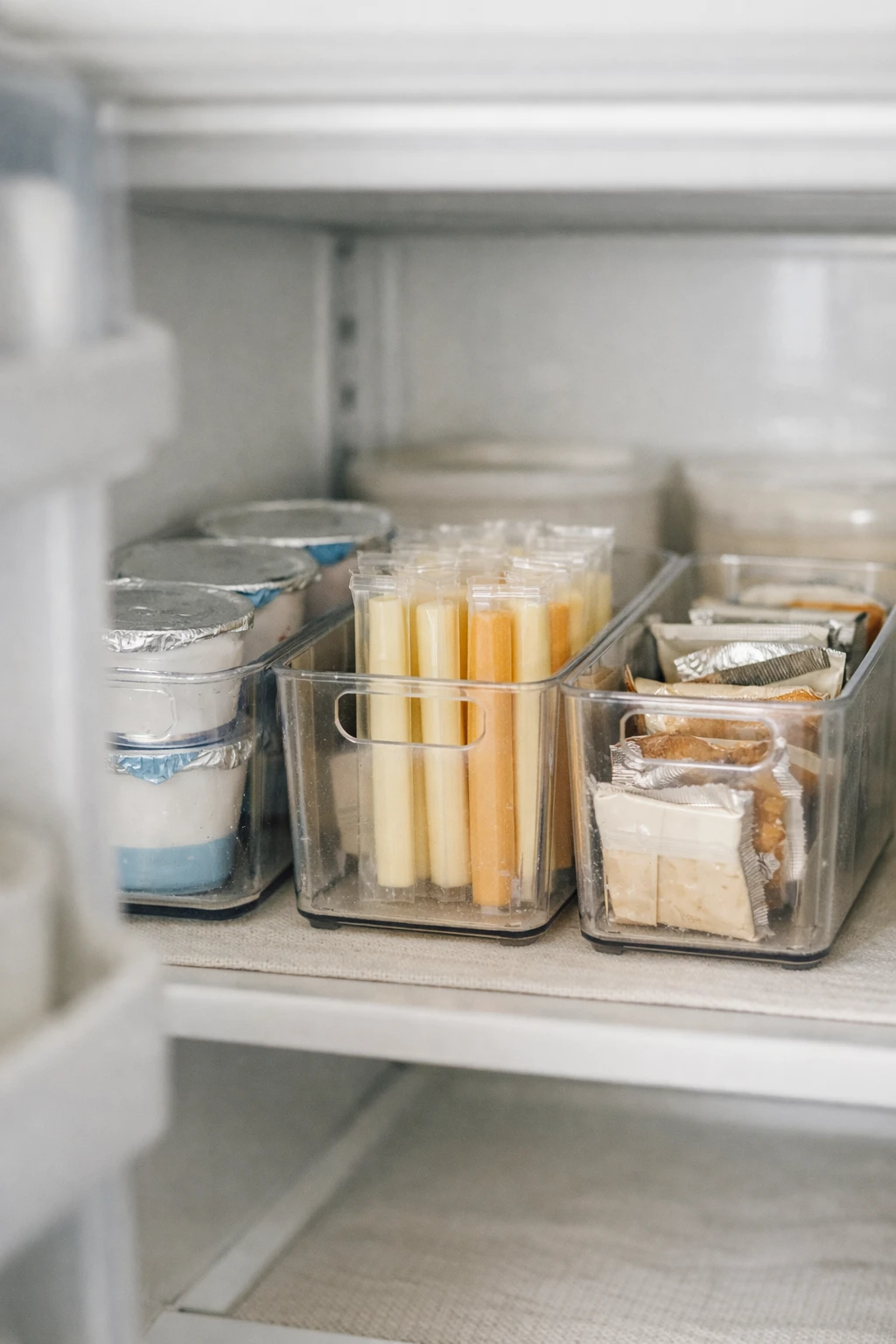 Refrigerator shelf organized with clear bins holding yogurt cups and snack packs